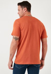 Kortærmet t-shirt i brændt orange, lavet af blødt stof, har rund hals og lige kant, med synlige tatoveringer på armen.
