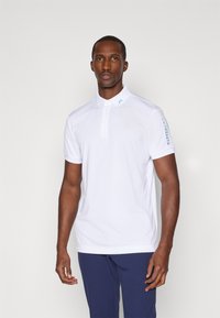 Hvit polo skjorte laget av glatt stoff, med klassisk krage, tre-knappers knappestolpe, og lyseblå logo på venstre skulder.