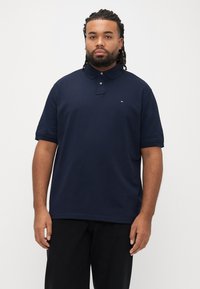 Marineblå polo med krave, korte ærmer, to knapper og et lille logo på brystet; lavet af bomuldslignende stof med glat tekstur.