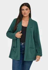 Moteris dėvi tamsiai žalią atvirą švarką su priekinėmis kišenėmis, juoda viršūne ir mėlynais džinsais, atsistojusi tiesiai ir viena ranka laiko švarką.