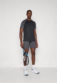 Homme portant un t-shirt et un short de sport noir et gris, tenant une raquette de tennis bleu et noir, avec des chaussettes blanches et des baskets, se tenant contre un mur blanc.