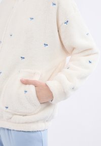 Kremowa polarowa kurtka z haftem w kształcie jasnoniebieskiej kokardy, z zapięciem na zamek błyskawiczny i przednimi kieszeniami. Miękka faktura i luźny krój.