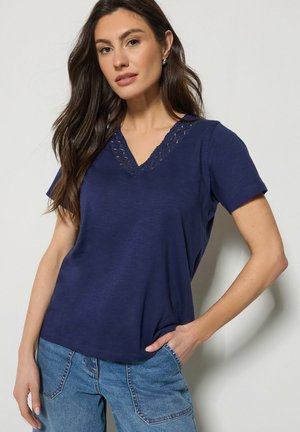 Vrouw met lang donker haar, gekleed in een marineblauw V-hals shirt met kanten rand en lichtblauwe spijkerbroek, poserend tegen een lichte achtergrond.