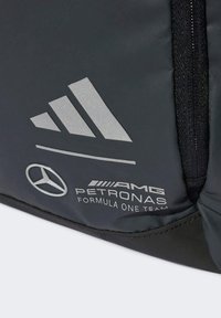 Crna ruksak s srebrnim logotipima, s brendiranjem Adidas i Mercedes AMG Petronas. Glatka tkanina, pravokutnog oblika, s čvrstim patentnim zatvaračima.