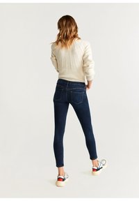 Jeans oscuros ajustados con una cintura alta y piernas estrechas, acompañados de un suéter crema y zapatillas de color claro. Textura acanalada en el suéter.