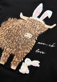 Černá tkanina obsahuje ilustraci v karikaturním stylu s texturovanou hnědou krávou s králičími ušima a vedle ní malého bílého králíka, text zní "moo-ch lásky."