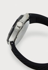 Montre avec un bracelet en caoutchouc noir, un boîtier en acier inoxydable, une couronne et un fond argenté. Le design est angulaire avec un bracelet texturé.