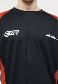 Μαύρη αθλητική φανέλα με κόκκινες πλαϊνές φάσες, λαιμόκοψη τύπου crew και λευκά, φλογερά λογότυπα στο στήθος, φορεμένη από ένα άτομο που φαίνεται από τον λαιμό έως το μέσο του κορμού.