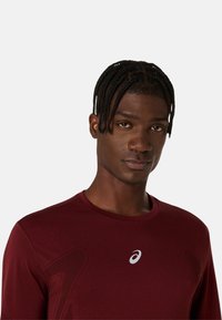 Bordeauxfarbendes Sportshirt mit Rundhalsausschnitt, strukturiertem Stoff und reflektierendem Logo auf der Brust. Nahaufnahme des Gesichts eines Modells.