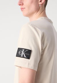 Beige tričko s krátkým rukávem, které na horním rameni zdobí černý obdélníkový patch s logem Calvin Klein Jeans, vyrobené z hladké bavlny.