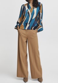 Blusa multicolore con un motivo astratto blu, marrone e crema, con scollatura a V e bottoni. Abbinata a pantaloni beige a gamba larga.
