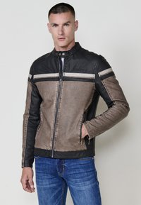 Uomo che indossa una giacca a zip marrone e nera con strisce sul petto e sulle maniche, abbinata a un paio di jeans blu, in piedi contro uno sfondo neutro.