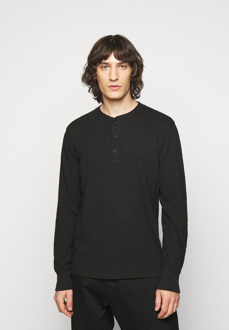 Black long-sleeve henley shirt featuring a rounded neckline, three-button placket, and a textured fabric. Styled with slim-fit black pants.
