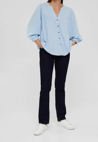 Blouse bleu clair avec un décolleté en V, des détails plissés et une fermeture à boutons. Associée avec un jean noir et des baskets blanches, présente une coupe décontractée.