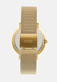 Guld mesh rem ur med rund skive, minimalistisk design, og "GUESS" gravert på låsen. Klar glasoverflade på urskiven.