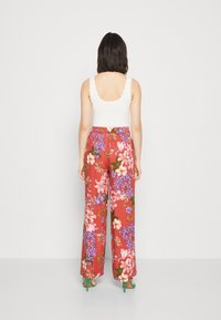 Witte tanktop met golfranden, gecombineerd met een wijde bloemenbroek in rood, met roze en blauwe bloemen. Zichtbare groene hoge hakken.