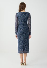 Abito a fantasia navy con maniche lunghe e trasparenti, caratterizzato da puntini colorati, una silhouette aderente e una lunghezza fino al polpaccio. Sono visibili sandali con tacco alto.