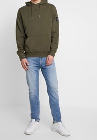 Olijfgroene hoodie met capuchon, kangoeroezak en logopatch, gecombineerd met lichtblauwe spijkerbroek en witte sneakers. Eenvoudig, casual ontwerp.