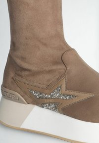 Stivaletto alla caviglia in suede marrone con un accento a stella luccicante, suola bianca e dettaglio del logo impresso. Texture liscia e forma arrotondata.