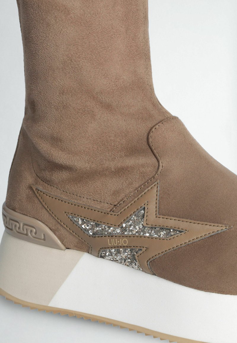 Bottines en daim marron avec un accent étoile pailleté, semelle blanche et logo embossé. Texture lisse et forme arrondie.