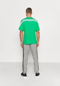 Camiseta verde de manga corta con tres franjas blancas en la parte posterior, combinada con pantalones de chándal grises que tienen franjas laterales negras y zapatillas negras.