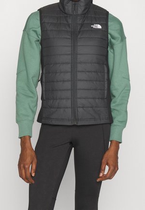 Person, die eine schwarze gesteppte North Face Weste über einem grünen Langarm-Sweatshirt und schwarze Hosen trägt, steht mit entspannten Armen.