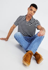 Camisa polo a rayas blancas y negras, jeans azul claro y zapatos de gamuza marrón con cordones. Modelo sentado con las piernas cruzadas sobre un fondo blanco.