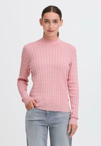 Maglione a maglia rosa chiaro con collo alto, polsini a coste e un motivo a treccia testurizzato, abbinato a jeans grigi chiari.