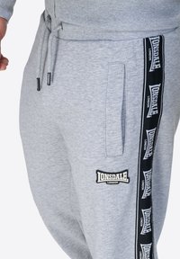 Pantalones de chándal grises hechos de algodón suave, con bolsillos laterales, que presentan una franja de logo negra a lo largo de la pierna y un parche con el logo en la parte frontal.