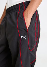 Zwarte sportbroek met rode zijspecificaties, een elastische tailleband en een grijze Puma-logo op het linkerpijp. Voorzien van zijzakken en een losse pasvorm.