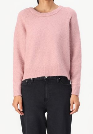 Femme portant un pull doux à manches longues rose pâle avec des poignets et un ourlet côtelés, associé à un jean noir taille haute.