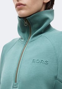 Κοντινό πλάνο ενός ατόμου που φοράει μια μπλούζα με φερμουάρ σε τιρκουάζ χρώμα, με ψηλό γιακά και ανάγλυφο λογότυπο "BORG" στο στήθος.