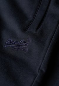 Maglione blu navy scuro con una consistenza morbida, caratterizzato da un logo "SuperDry" ricamato in viola e dettagli di cucitura sottili.