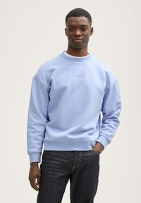 Lyseblå sweatshirt i blødt stof med ribbetrimmede manchetter og kant, med et hvidt logo trykt på brystet, parret med mørke denimjeans.