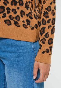 Pull en motif léopard de couleur brun doré avec des accents noirs, fabriqué à partir d'un matériau tricot doux. Présente un ourlet et des poignets côtelés.