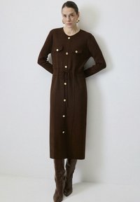 Robe tricotée marron avec un devant à boutons, deux poches poitrine et une taille à cordon ; assortie à des bottes marron jusqu'aux genoux.
