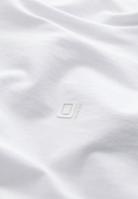 Lähikuva valkoisesta kankaasta, jossa on hienovaraisesti kohokuvioitu logo, jossa näkyy kirjain O ja sana "OUTDOOR" pienellä painatuksella.