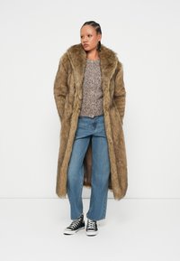 Μακρύ παλτό από φελλό σε ανοιχτό καφέ, με μεγάλο γιακά, φορεμένο πάνω από έναν γκρι υφασμάτινο σέντερ και μπλε φαρδιά παντελόνια με μαύρα αθλητικά παπούτσια.