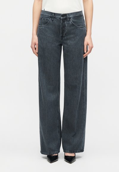 Femme portant un jean gris foncé taille haute à jambes larges, associé à des talons noirs à bout pointu, debout devant un fond blanc uni.