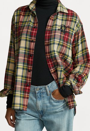 Personne portant une chemise à carreaux rouge, jaune et verte par-dessus un col roulé noir, avec la main dans la poche d'un jean bleu clair.