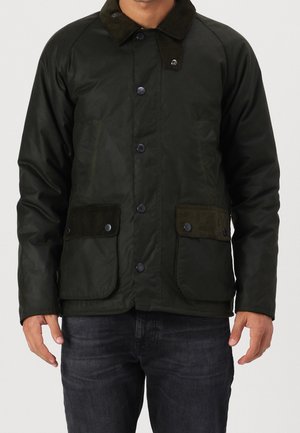 Man draagt een donkergroen jasje met knopen, twee klepzakken aan de voorkant en een corduroy kraag, gecombineerd met een donkere spijkerbroek.