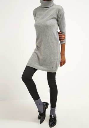 Vrouw draagt een grijze, lange coltrui-jurk met lange mouwen, zwarte legging, grijze sokken en zwarte loafers met kwastjes.