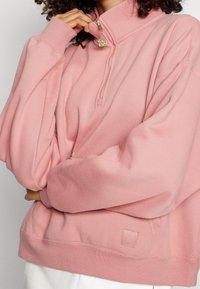 Sudadera de color rosa claro con cuello alto, detalle de cremallera y un acabado texturizado. Presenta un pequeño logo en relieve en el dobladillo.