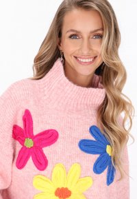 Rosa gestrickter Pullover mit hohem Rippsaumkragen, der große gestickte Blumen in Pink, Blau und Gelb zeigt. Weiches, strukturiertes Material.