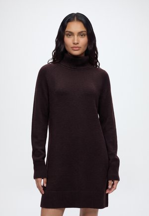 Un mannequin se tient devant un fond uni et clair, vêtu d’une robe en maille marron foncé à manches longues, avec un col montant côtelé et des poignets côtelés.