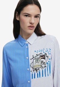 Camicia a righe con bottoni in tessuto blu e bianco. Il lato sinistro presenta sottili strisce verticali; il lato destro mostra un design grafico e del testo.