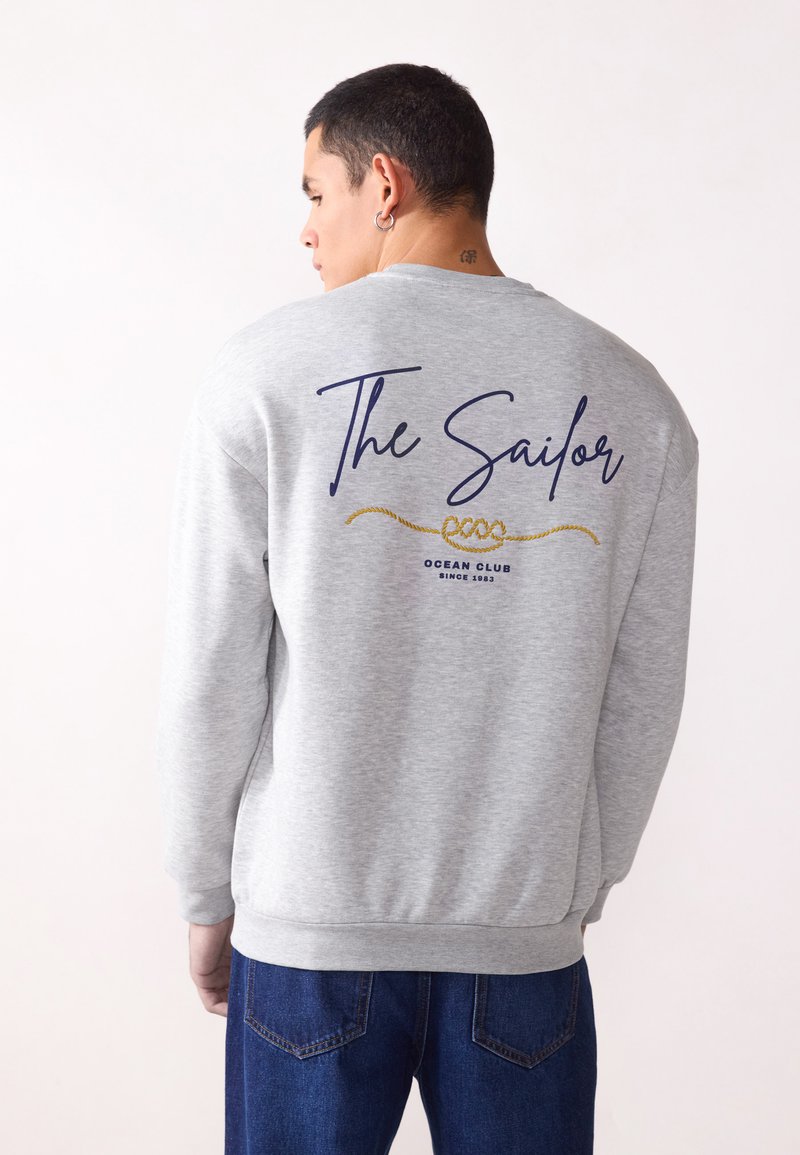 Grå hettegenser med "The Sailor" trykket i marineblått, med et gulltau-detalj. Ribbestrikkede mansjetter og hem, brukt sammen med blå denimbukser.