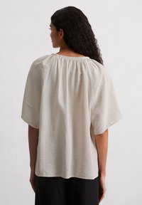 Hellebeige Bluse mit gerafftem Ausschnitt, kurzen Ärmeln, lockerer Passform und strukturiertem Stoff. Keine sichtbaren Muster oder Zubehördetails.
