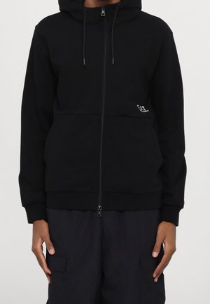 Person trägt einen schwarzen Zip-Hoodie mit kleinem weißem Logo auf der linken Seite und schwarze Cargohosen, steht vor einem schlichten Hintergrund.