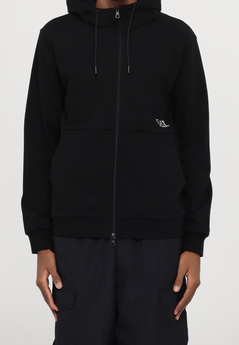 Personne portant un sweat à capuche zippé noir avec un petit logo blanc sur le côté gauche et un pantalon cargo noir, debout devant un fond uni.